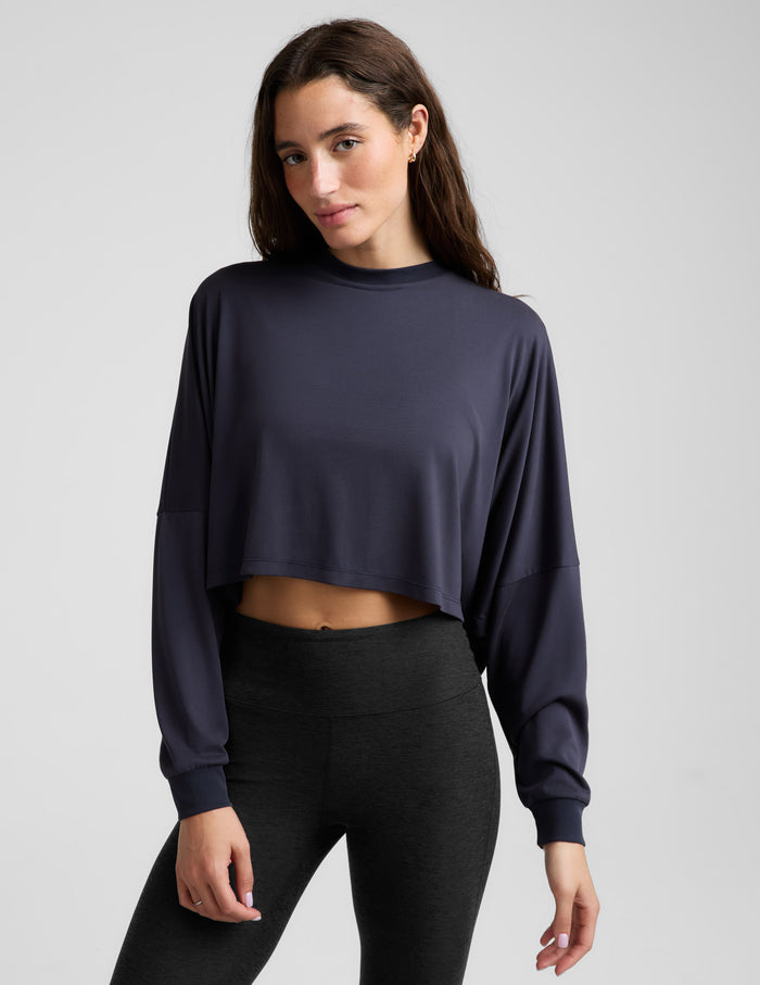 A woman modeling a dark navy blue long-sleeve crop top, paired with black leggings, against a solid light gray background.