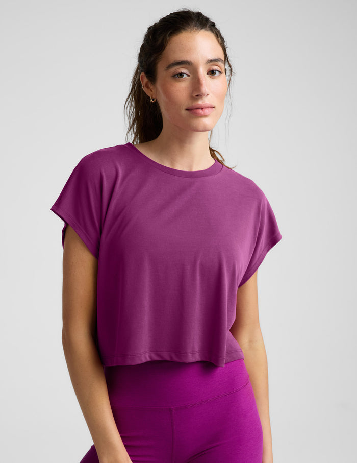 A woman wearing a short-sleeve cropped athletic top and matching biker shorts in a solid magenta-purple color, photographed against a light gray studio background.