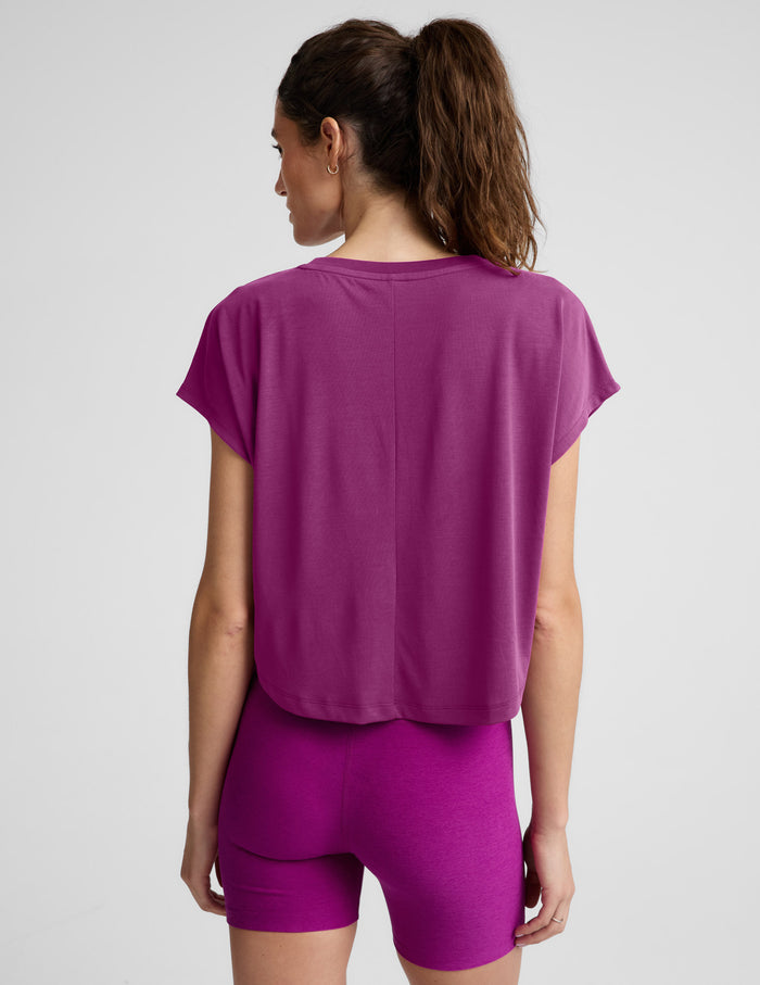 A woman wearing a short-sleeve cropped athletic top and matching biker shorts in a solid magenta-purple color, photographed against a light gray studio background.