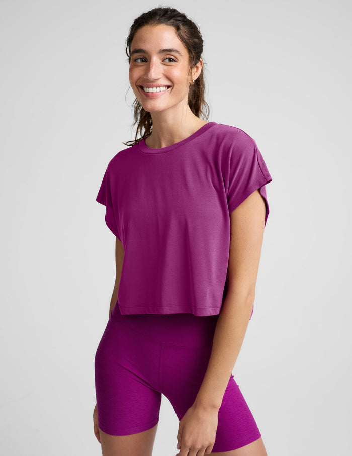 A woman wearing a short-sleeve cropped athletic top and matching biker shorts in a solid magenta-purple color, photographed against a light gray studio background.