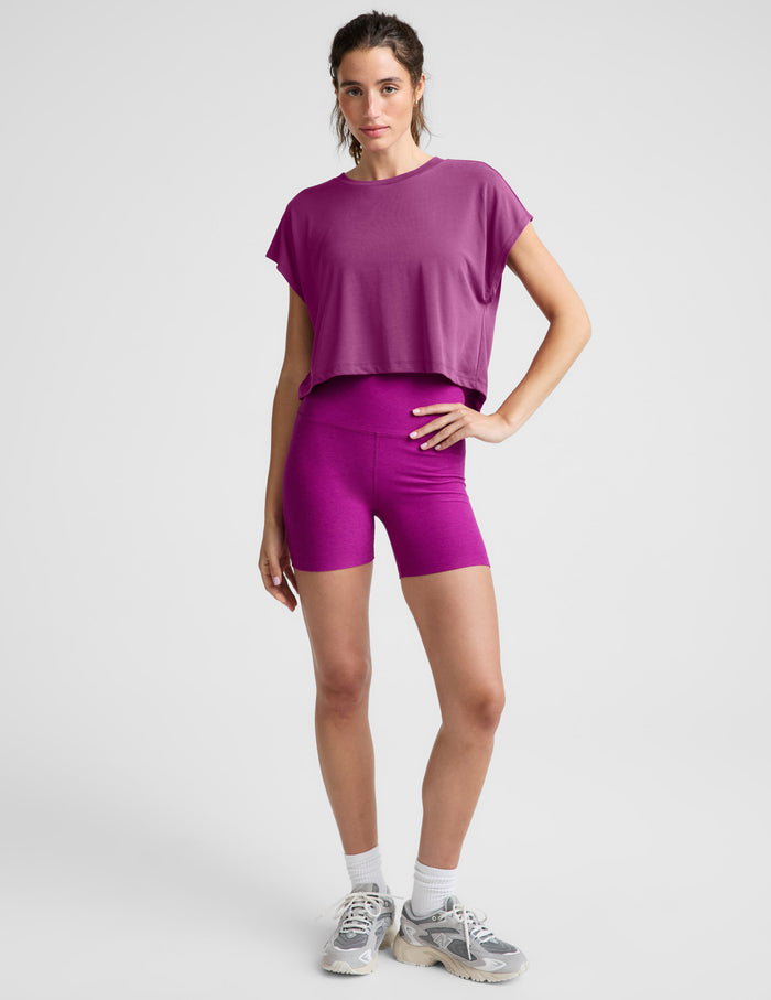 A woman wearing a short-sleeve cropped athletic top and matching biker shorts in a solid magenta-purple color, photographed against a light gray studio background.