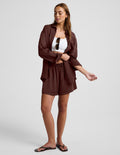 A woman modeling a matching brown lounge set consisting of a long-sleeve button-up shirt and shorts, styled over a white crop top and black flip-flops against a light gray studio background.