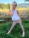 Toddler wearing a white tank top and gingham printed leggings standing on grass.