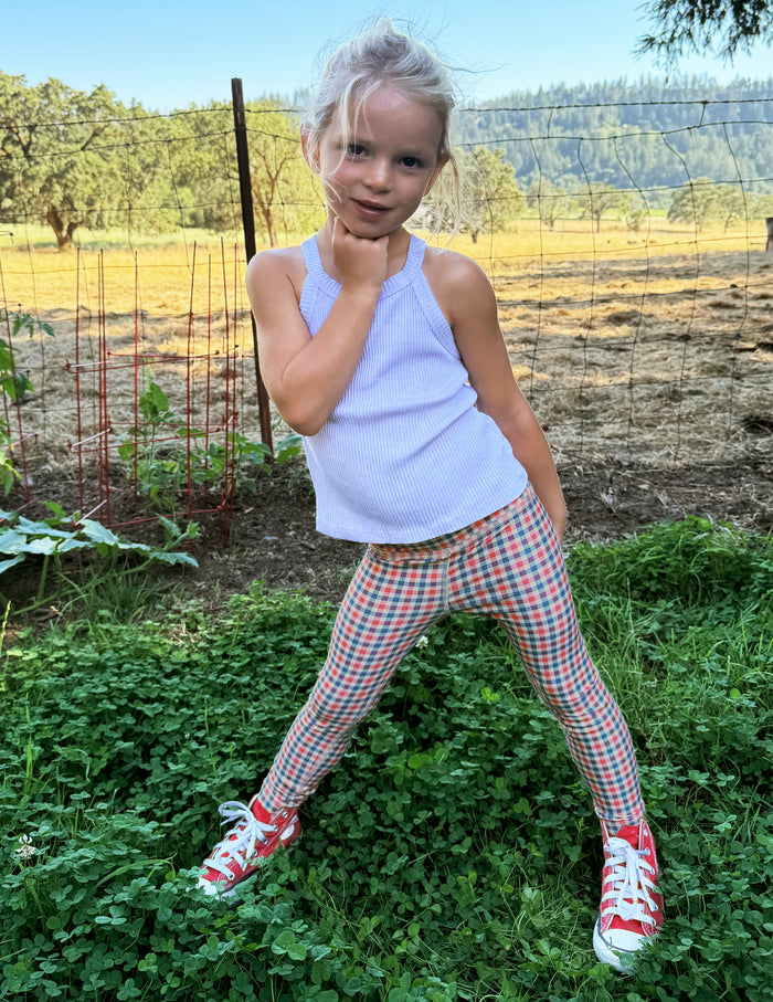 Toddler wearing a white tank top and gingham printed leggings standing on grass.