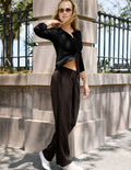 A woman wearing a black long-sleeve button-up shirt and dark brown wide-leg pants, posing outdoors in front of a stone wall and black metal fence.