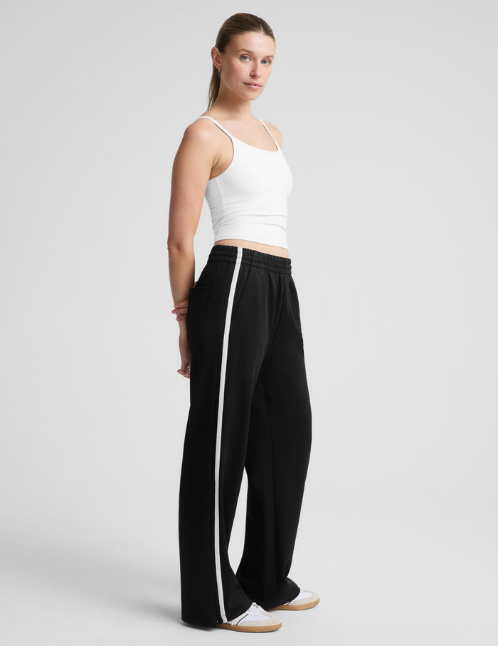A woman modeling a white spaghetti-strap tank top and black wide-leg track pants against a light gray studio background.