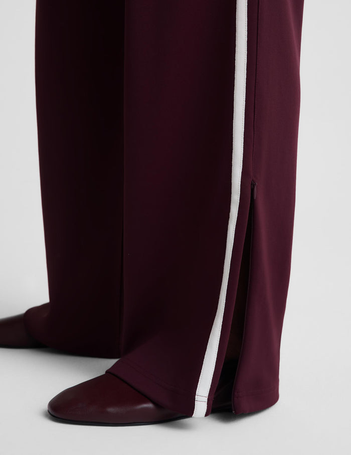 Close-up of dark burgundy wide-leg trousers with a vertical white stripe down the side and a front slit, paired with matching burgundy pointed flats on a light gray studio background.