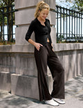A woman posing outdoors in a black long-sleeve button-up top and dark brown wide-leg trousers, leaning against a stone column next to a black metal fence.