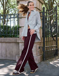 A woman posing outdoors in a stylish outfit featuring a light gray and white pinstriped shirt, wide-leg trousers with a white side stripe, and dark burgundy sneakers.