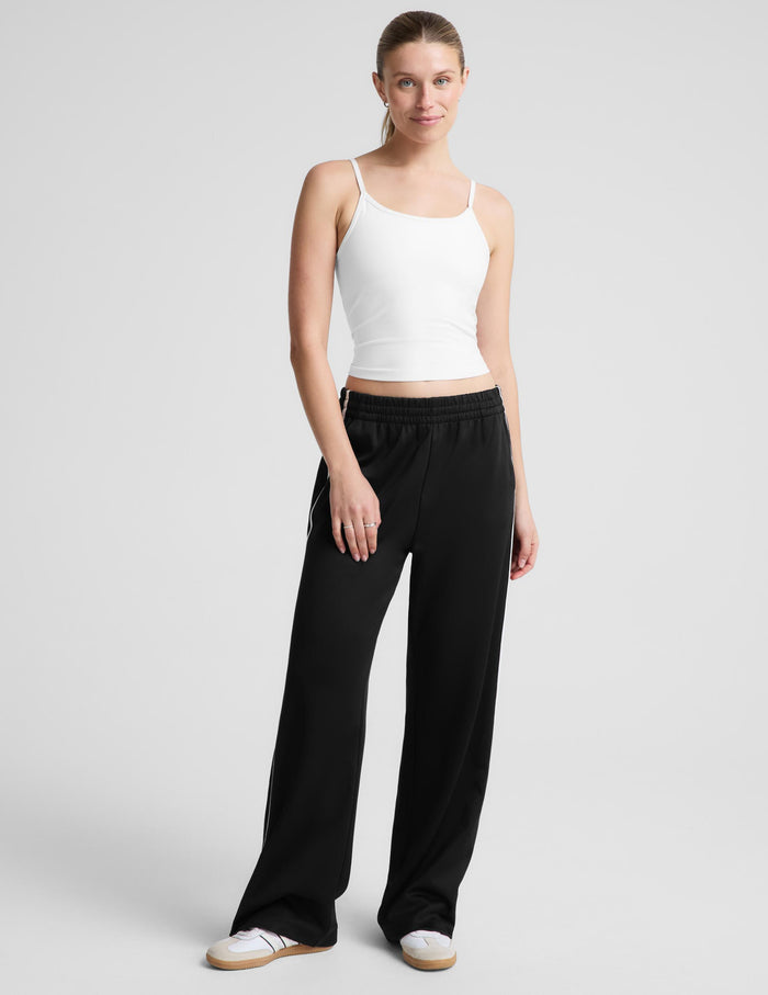 A woman modeling a white spaghetti-strap tank top and black wide-leg track pants against a light gray studio background.