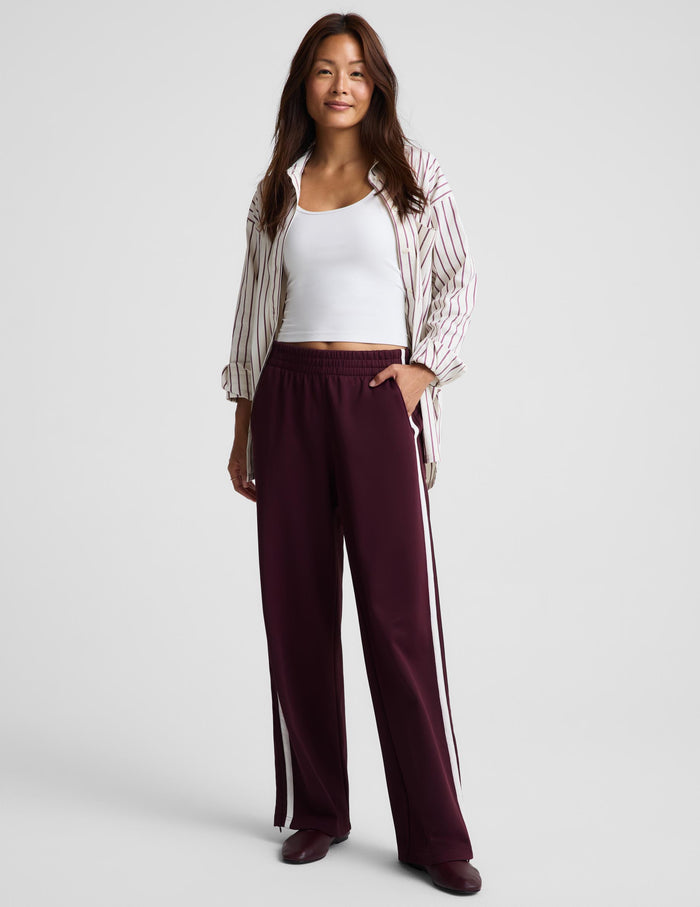 A woman modeling a pair of burgundy wide-leg track pants with white side stripes, styled with a white tank top, a striped button-up shirt, and dark brown shoes against a light gray studio background.