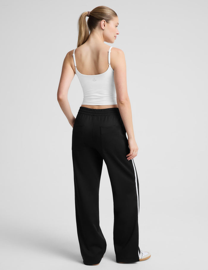 A woman modeling a white spaghetti-strap tank top and black wide-leg track pants against a light gray studio background.
