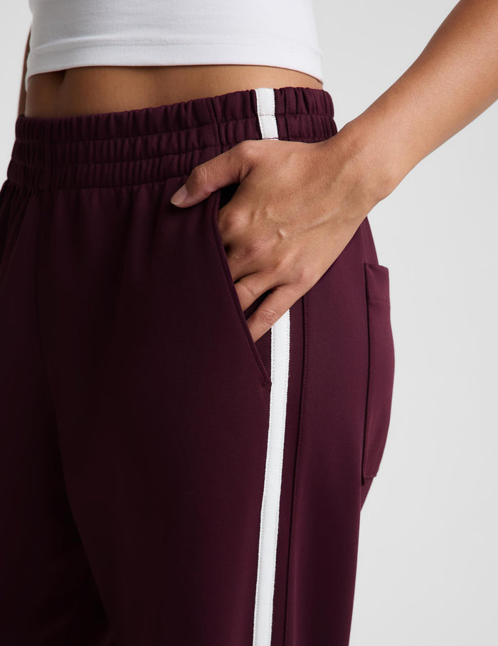 Close-up of a person wearing burgundy athletic pants with a white side stripe and white waistband detail, shown against a light gray background.