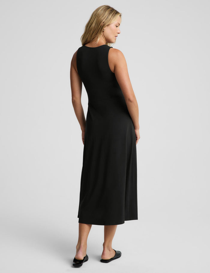 A woman modeling a sleeveless black midi dress from the back, standing against a light gray studio background.