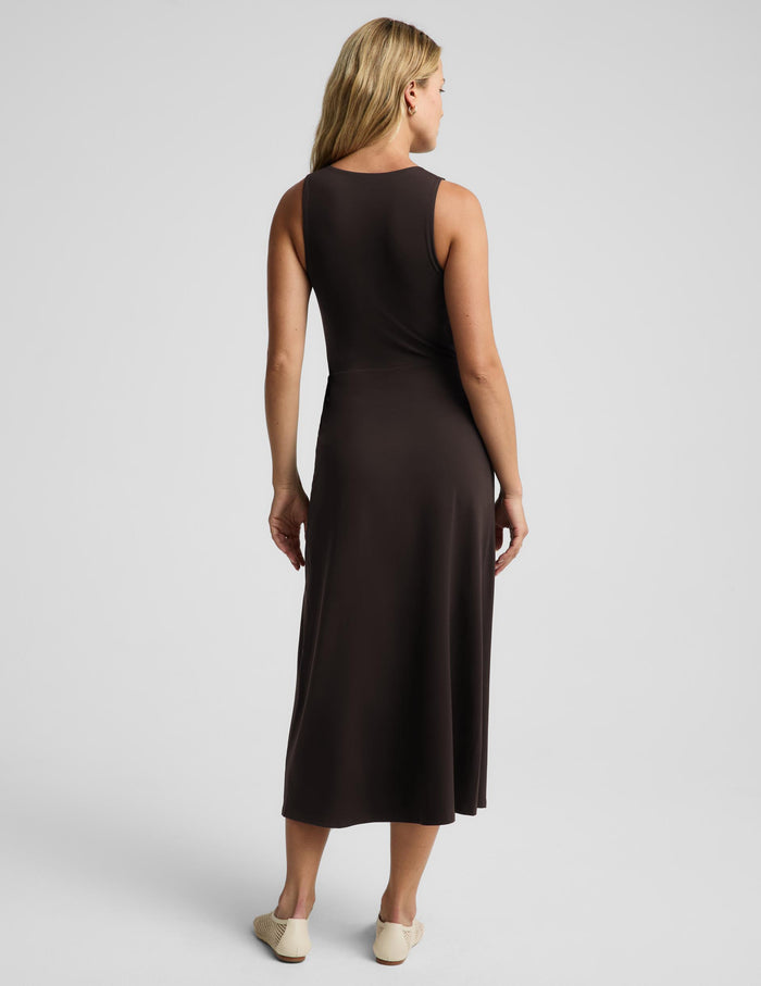 A woman viewed from the back wearing a sleeveless dark brown midi dress and light beige flats, standing against a light gray studio background.