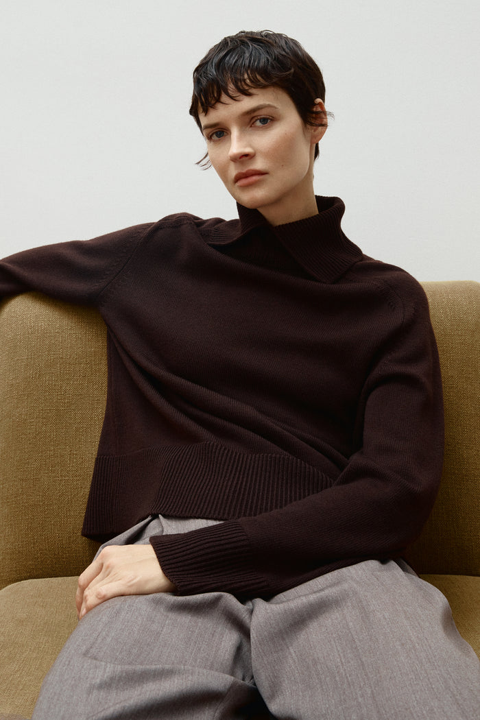 Person wearing a brown Merino Wool Boxy with special High Neck  on a beige couch.