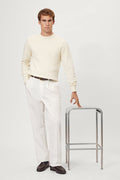 Man in a wool white sweater and white pants holding a metal stool on a plain background