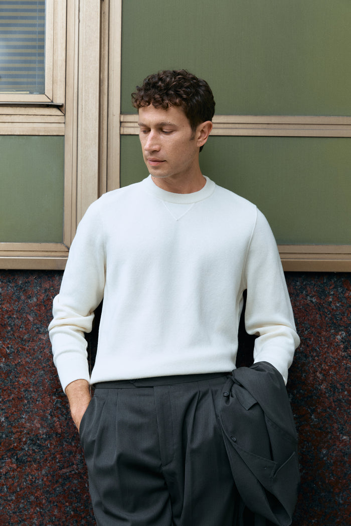 Man wearing a white wool sweater and dark pants against a green wall.