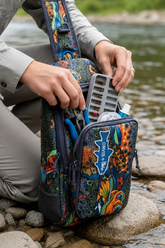 Person organizing items in a colorful fishing tackle bag by a river.