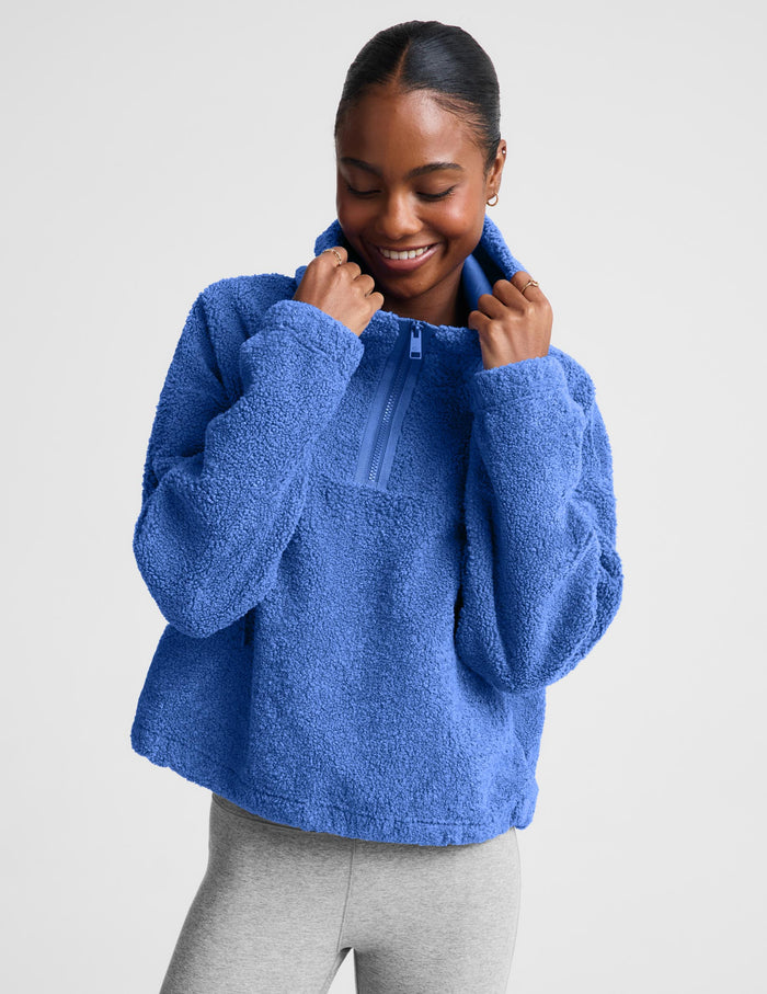 A woman wearing a textured blue fleece quarter-zip pullover sweater and gray leggings, posing against a light gray studio background.