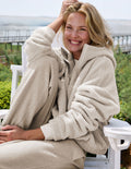 A smiling woman sits in a white outdoor chair wearing a cozy beige sherpa zip-up hoodie and matching sweatpants, with a blurred garden and water background.
