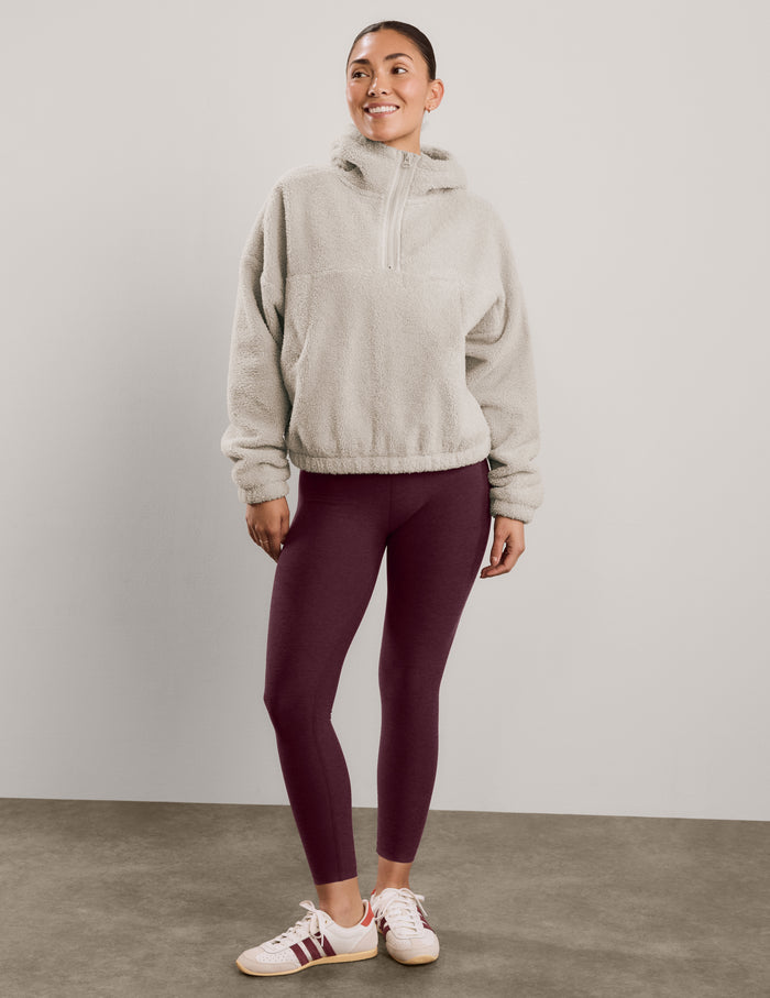 A woman modeling a light beige sherpa fleece half-zip pullover sweatshirt with a high collar, paired with dark burgundy leggings, against a solid light gray studio background.