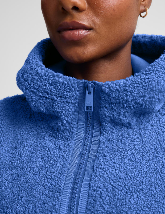A woman wearing a textured blue fleece quarter-zip pullover sweater and gray leggings, posing against a light gray studio background.