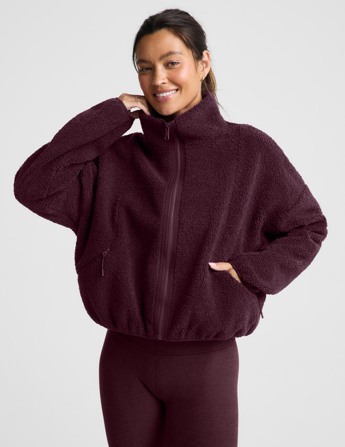 A woman wearing a dark burgundy boucle zip-up jacket and matching leggings, posing against a light gray studio background.