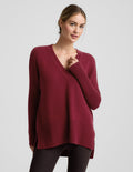 Woman wearing a red v-neck long length sweater and brown leggings against a plain background.