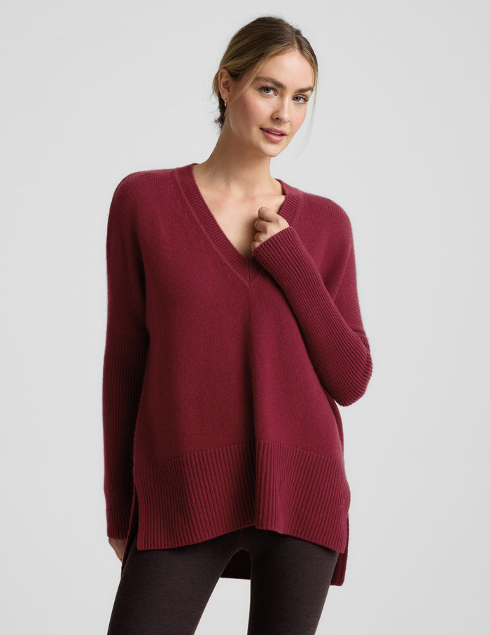 Woman wearing a red v-neck long length sweater and brown leggings against a plain background.