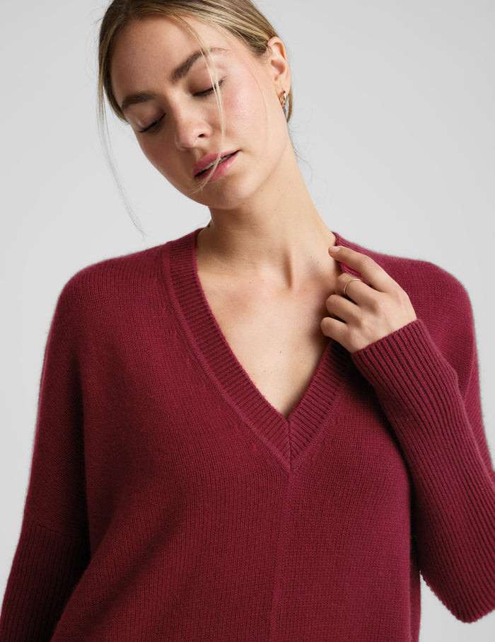 Woman wearing a red v-neck long length sweater and brown leggings against a plain background.
