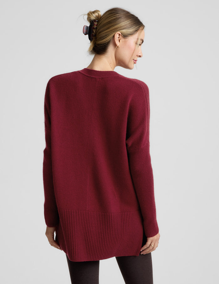 Woman wearing a red v-neck long length sweater and brown leggings against a plain background.