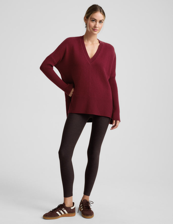 Woman wearing a red v-neck long length sweater and brown leggings against a plain background.