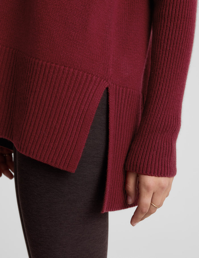 Woman wearing a red v-neck long length sweater and brown leggings against a plain background.